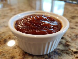 Close-up of rich, dark homemade bbq sauce served in a small white ramekin on a granite countertop.