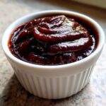 Close-up of thick, dark, glossy homemade bbq sauce swirled in a small white ramekin dish.