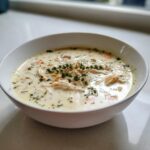 A close-up of a white bowl filled with creamy homemade cream of chicken soup, garnished with shredded chicken and fresh thyme.