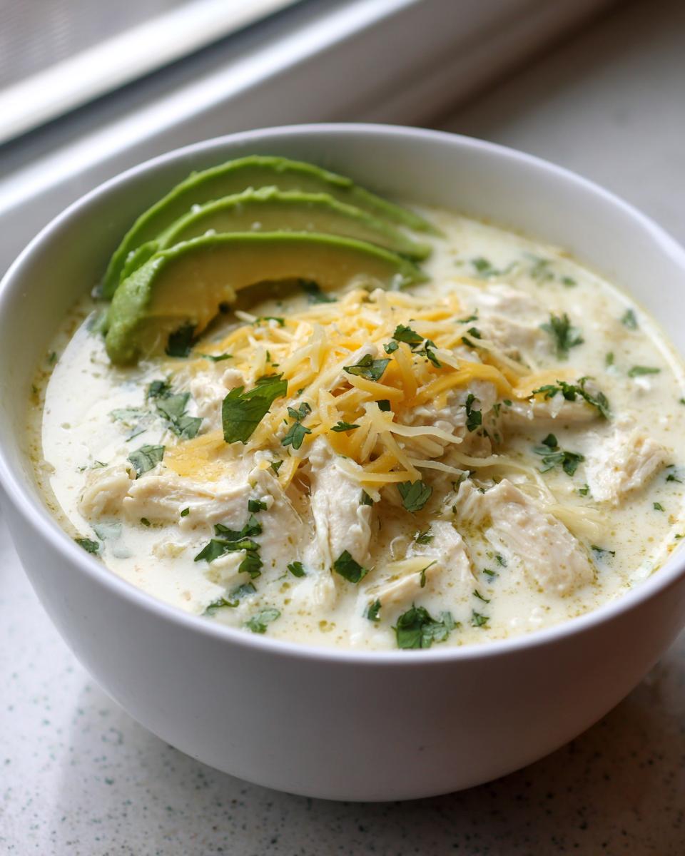 A white bowl filled with creamy keto white chicken chili, topped with shredded cheese, cilantro, and sliced avocado.