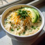 A close-up of a bowl of creamy keto white chicken chili topped with shredded cheese, cilantro, and avocado slices.