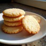 A stack of four golden brown sugar cookies with one leaning against the stack on a white plate.