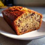 Close-up of a freshly baked, moist banana bread recipe loaf with visible chocolate chips on a white plate.