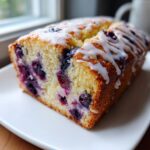 A close-up of a slice of moist blueberry bread topped with a white vanilla glaze, showing abundant blueberries inside.