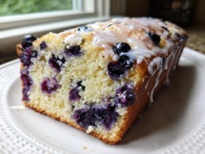 Close-up of a moist slice of blueberry bread topped with a sweet white glaze, showing vibrant blueberries inside.