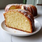 A close-up of a moist slice of olive oil cake on a white plate, drizzled with white lemon glaze.