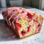 Close-up of a moist loaf of strawberry bread studded with fresh strawberries and drizzled with pink glaze.