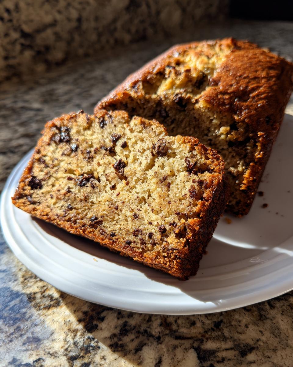 A close-up of a moist slice of vegan banana bread studded with chocolate chips, resting on a white plate.