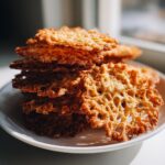 A stack of golden brown, delicate oatmeal lace cookies showing their intricate, lacy texture on a white plate.