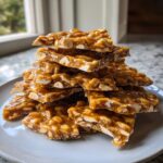 A stack of broken, golden-brown pieces of homemade peanut brittle recipe piled high on a light gray plate.
