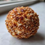 A perfectly round cheese ball recipe coated entirely in chopped pecans and spices, sitting on a white surface.