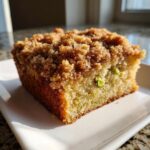 A close-up, sunlit slice of pistachio coffee cake featuring a thick, sugary brown crumb topping and visible green pistachio pieces.
