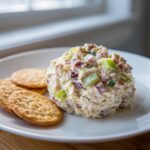 A scoop of creamy ham salad recipe mixed with celery and red ingredients, served with round crackers on a white plate.