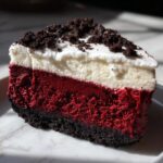 A perfect slice of red velvet cheesecake showing three distinct layers: dark chocolate crust, vibrant red filling, and white cream cheese topping with chocolate crumbs.