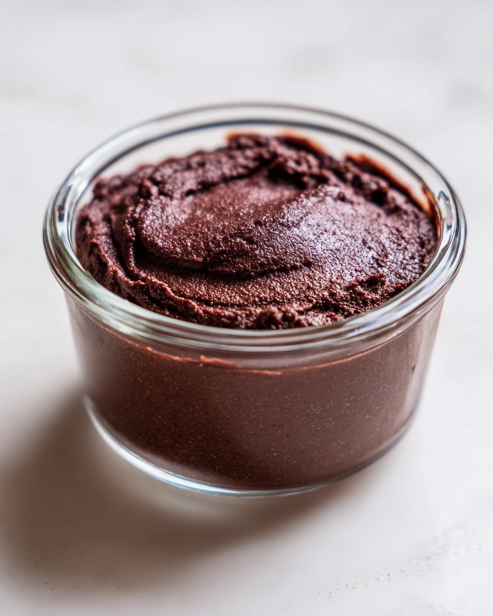 A close-up of thick, dark chocolate pudding served in a small glass ramekin on a white surface.