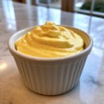 A close-up of thick, vibrant yellow hollandaise sauce swirled high in a white, fluted ramekin.