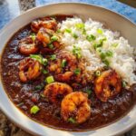A close-up of a bowl of dark, rich shrimp etouffee next to a mound of white rice, garnished with green onions.