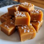 A close-up stack of homemade, soft, square caramels topped generously with flaky sea salt.