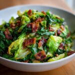 Close-up of a white bowl filled with sautéed green cabbage recipe pieces topped generously with crispy bacon bits.