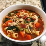 Close-up of a steaming bowl of Sicilian chicken soup filled with shredded chicken, carrots, celery, tomatoes, and pasta.
