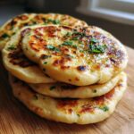A stack of four freshly cooked, golden-brown naan bread recipe rounds brushed with butter and topped with chopped parsley.