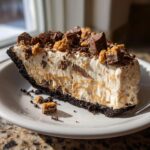 A thick slice of no-bake butterfinger pie with a dark crust, creamy filling, and topped with chopped candy pieces.