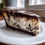A decadent slice of oreo cheesecake with a thick chocolate crust, creamy filling studded with cookie pieces, and topped with glossy ganache.