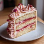 A perfect slice of layered rose cake featuring vanilla sponge, pink jam filling, and piped pink frosting topped with dried rosebuds.