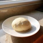 A smooth ball of light yellow fufu dough resting on a white plate, illuminated by sunlight.