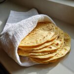 A stack of freshly made, soft corn tortillas wrapped partially in a white cloth, kept warm.