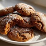 Close-up of several soft gingerbread cookies piled on a white plate, dusted with powdered sugar; one cookie is broken open showing the soft interior.