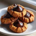 Close-up of several soft peanut butter blossom cookies stacked on a white plate, topped with melted chocolate kisses.