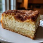 A close-up of a thick slice of sopapilla cheesecake showing a creamy white filling and a dark cinnamon-sugar crust.