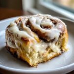 A close-up shot of a single, fluffy sourdough discard cinnamon roll covered in thick white icing on a white plate.