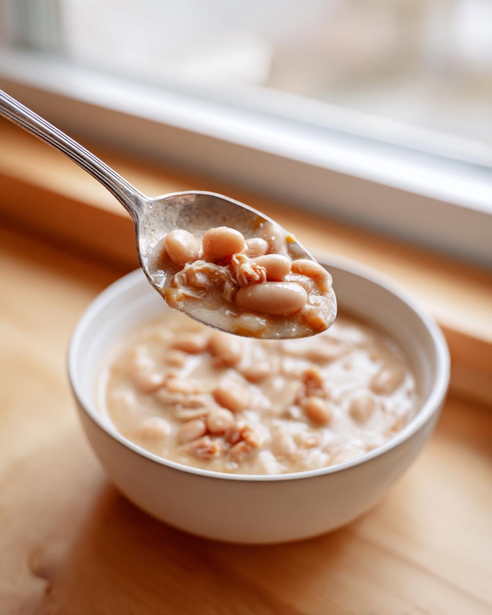 A spoonful of creamy instant pot white chicken chili, showing white beans and shredded chicken, lifted above a white bowl.