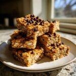 A stack of square no bake granola bars topped with mini chocolate chips, sitting on a light-colored plate.