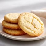 A stack of four golden brown, perfectly round soft sugar cookies resting on a white plate.
