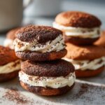 Close-up of stacked, decadent tiramisu cookies featuring dark chocolate wafers and thick vanilla cream filling, dusted with cocoa.