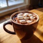 A steaming mug of rich homemade hot cocoa topped with fluffy white marshmallows, sitting on a wooden table near a window.