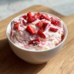 A close-up of a white bowl filled with creamy strawberry cheesecake dip, topped with fresh chopped strawberries.