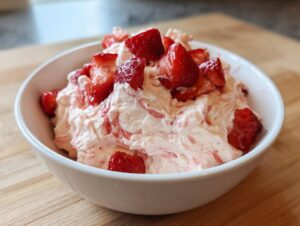 A white bowl filled with fluffy pink strawberry cheesecake dip, topped with fresh strawberry chunks.