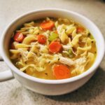 A close-up shot of a white bowl filled with steaming turkey noodle soup, featuring egg noodles, shredded turkey, and bright carrot slices.