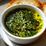 Close-up of a vibrant olive oil bread dip, rich with chopped green herbs, garlic, and red pepper flakes, served with crusty bread.