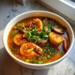 Close-up of a white bowl filled with spicy, orange broth tom yum soup, featuring large seasoned shrimp and sliced mushrooms, garnished with cilantro.