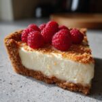 A close-up of a slice of white chocolate mousse tart with a thick graham cracker crust, topped with fresh raspberries.