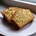 Three moist slices of zucchini banana bread, showing flecks of green zucchini, served on a white plate near a window.