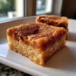 Two squares of soft, thick snickerdoodle bars topped with a rich cinnamon-sugar crust, served on a white plate.
