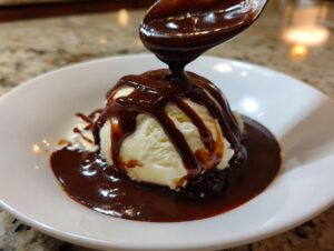 A spoonful of thick, glossy chocolate sauce is being poured over a scoop of vanilla ice cream in a white bowl.