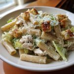 Close-up of a bowl of chicken caesar pasta salad featuring rigatoni, shredded chicken, romaine lettuce, and croutons.