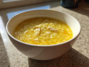 A close-up of a white bowl filled with bright yellow chicken lemon rice soup, featuring visible rice grains and shredded chicken.